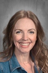 Portrait of a smiling woman with wavy light brown hair in a denim jacket, gray studio background.