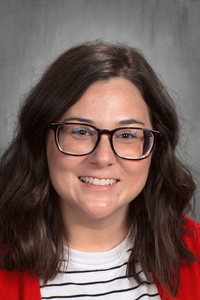 Smiling portrait of a woman with dark hair, glasses, red cardigan, and striped shirt against a gray backdrop.