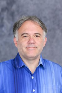 Portrait of a middle-aged man with gray hair in a blue striped button-down shirt, smiling against a gray backdrop.